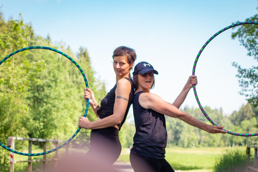Zwei Frauen stehen Rücken an Rücken und halten bunte Hula-Hoop-Reifen im Grünen.“